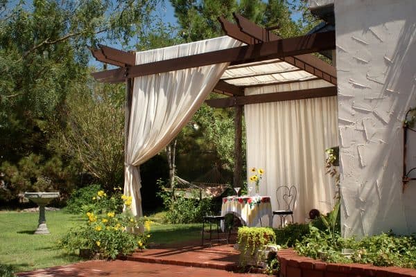 "A brick patio with wooden patio posts and a cover of canvas curtains. The back ground has green grass, a birdbath and hammock.", How To Keep Outdoor Curtains From Fading