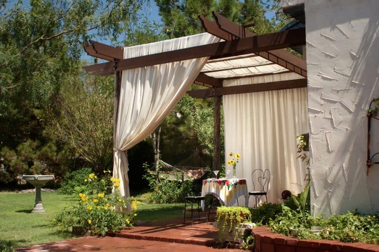 "A brick patio with wooden patio posts and a cover of canvas curtains. The back ground has green grass, a birdbath and hammock.", How To Keep Outdoor Curtains From Fading