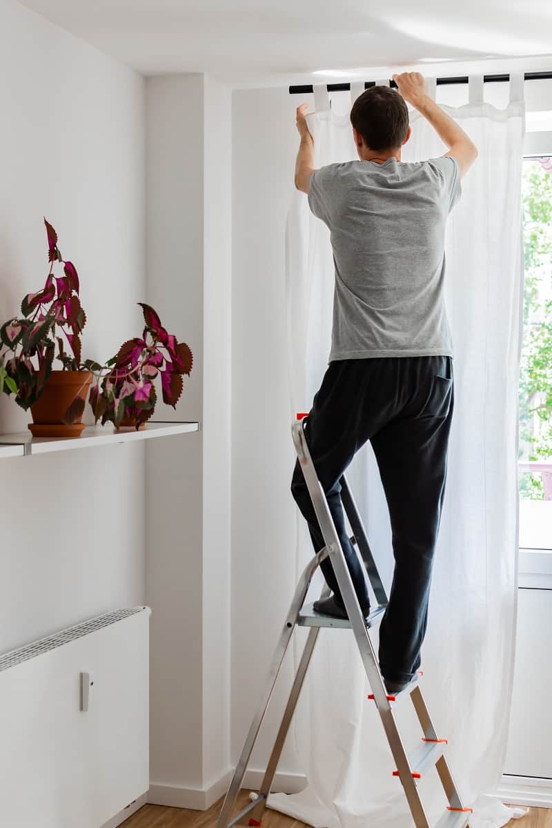 Man,Stands,On,A,Stepladder,Near,The,Window,And,Hangs - How High To Hang Curtains