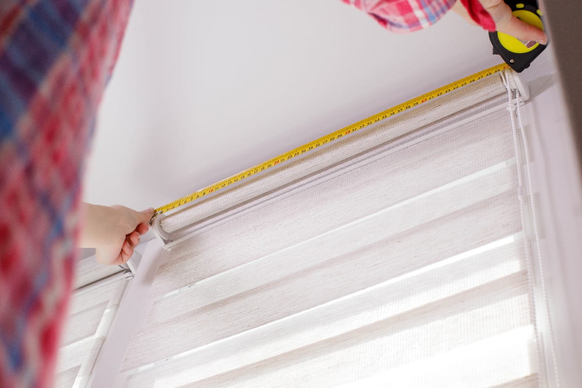 A handywoman with a tape measure installs white roller blinds indoors