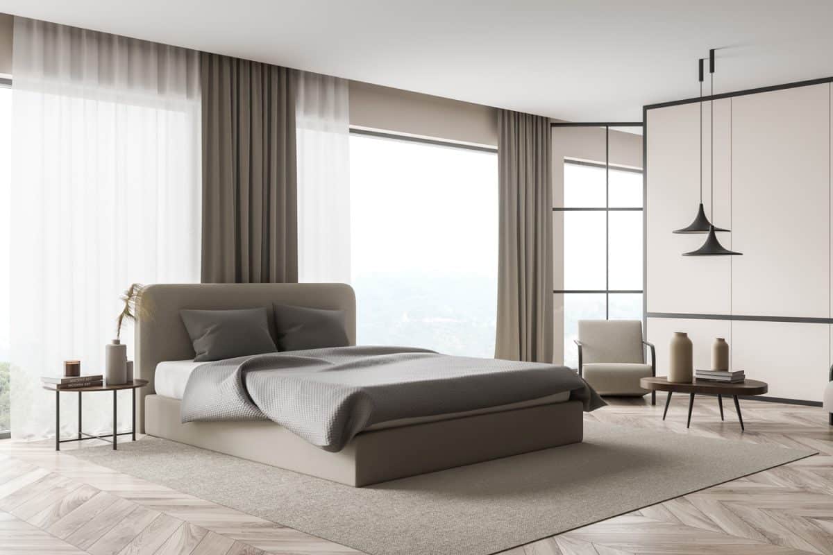 Corner view of the beige bedroom interior with two lamps over a seating area, panoramic windows with curtains, a bed and a parquet floor. A concept of modern trends in design.