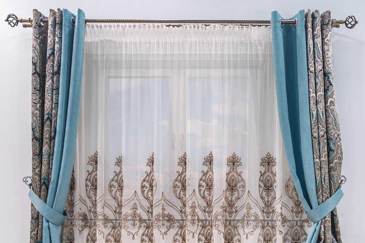Modern design of a small window. Combined curtain with eyelets, monochromatic turquoise fabric, tapestry with an ornament and a tulle with patterns hang on a round cornice.

