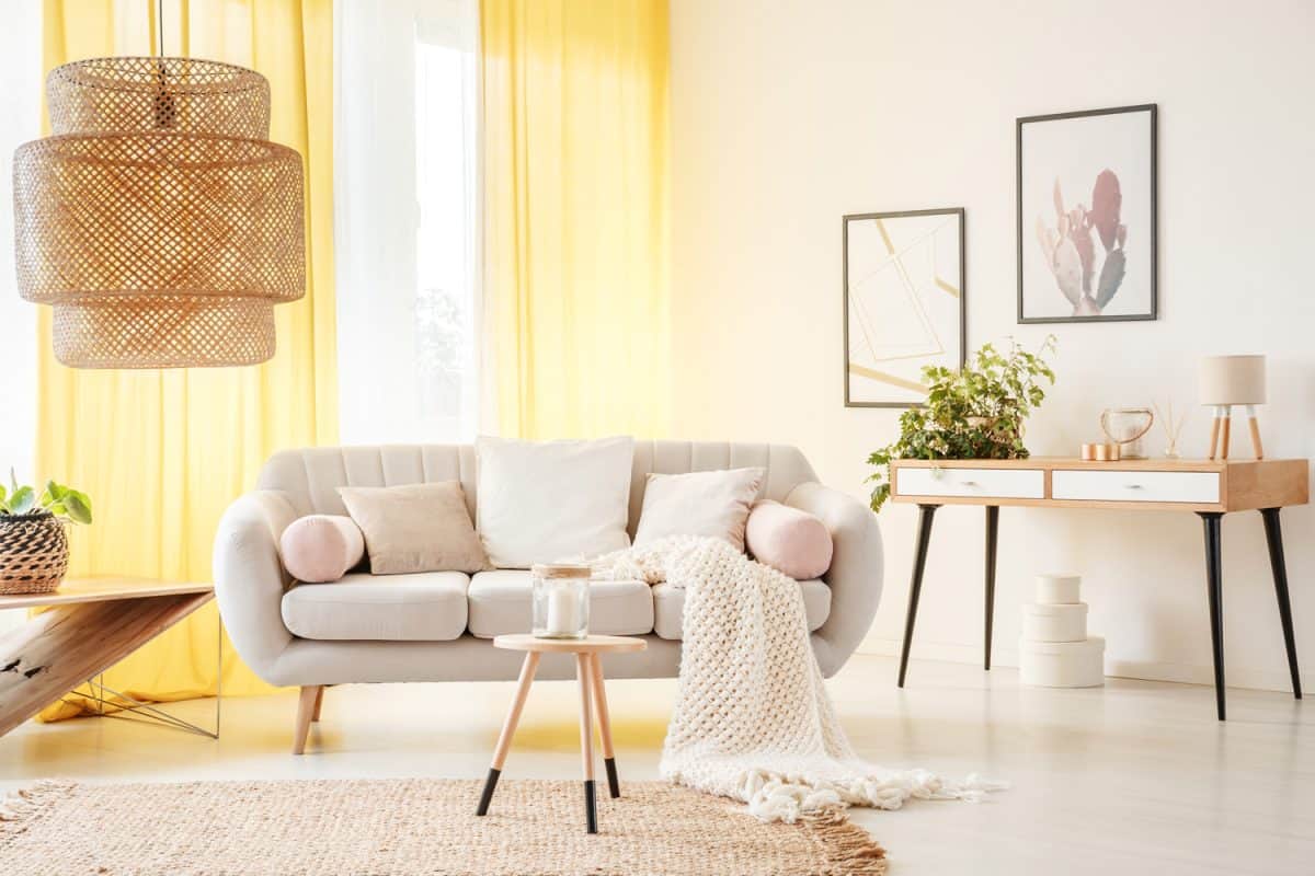 Rattan lamp and wooden stool on carpet in warm bohemian living room with yellow curtains and beige settee with white pillows
