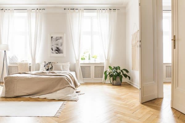Spacious and bright bedroom interior with beige decorations, hardwood floor and a book on the window sill seat - How To Hang Floor To Ceiling Curtains [11 Different Ways To Inspire You With Pictures]