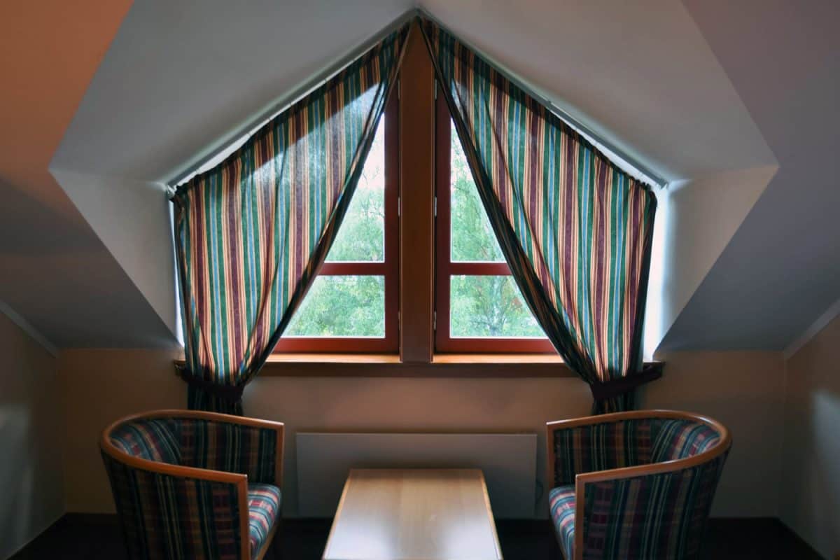 View of a non-standard window with curtains in the attic with asymmetric architecture, a table with two soft chairs

