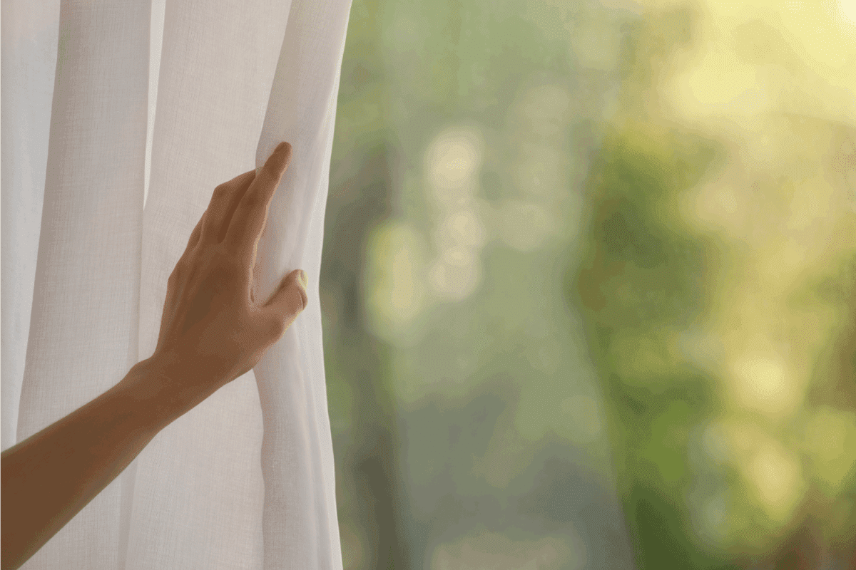 Woman opening white window curtains in room, closeup.