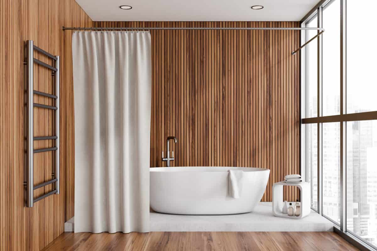 Bright bathroom interior with bathtub, panoramic window with city view, wooden walls, liquid soap, shower curtain and oak floor. Concept of hygienic and spa procedures.